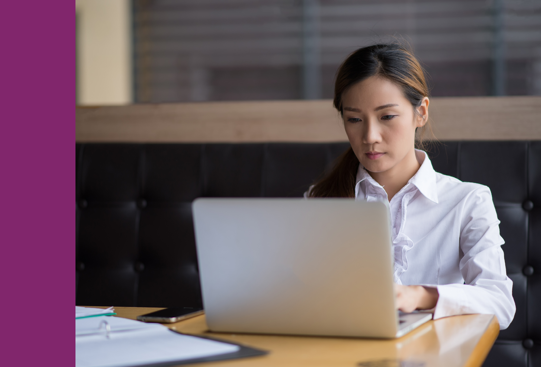 A woman in a white shirt sits at a table working on a laptop, with papers and a phone nearby.