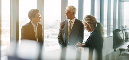 Three senior business colleagues standing in a boardroom in front of a window, having a discussion.