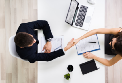 Two people sit across a desk in a modern office, shaking hands.