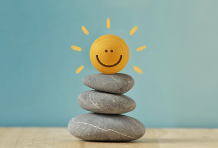 A minimalist scene of three smooth gray stones stacked on a wooden surface, topped with a small yellow ball painted with a smiling face. Simple sun rays are drawn around the ball, set against a soft blue background, evoking calm and positivity.