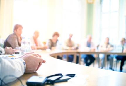  Locally Uploaded Image  This image was uploaded directly to AltText AI for processing. 	  People are seated around a curved conference table in a brightly lit room, with focus on hands in the foreground and blurred figures in the background. 