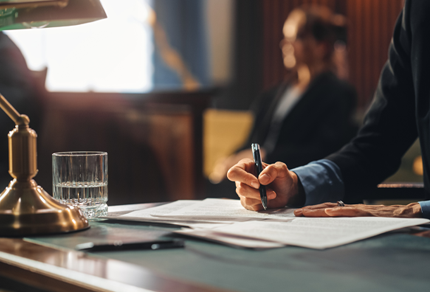 Person in professional attire writing on documents at a desk with a glass of water and a brass lamp in a courtroom or office setting.