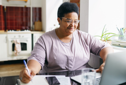 A woman in a white shirt sits at a table working on a laptop, with papers and a phone nearby.