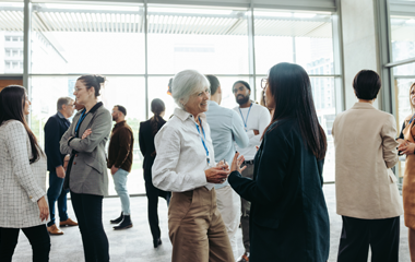 An older man with white hair, wearing a light button-down shirt and a lanyard, appears to be speaking and gesturing with his hands.