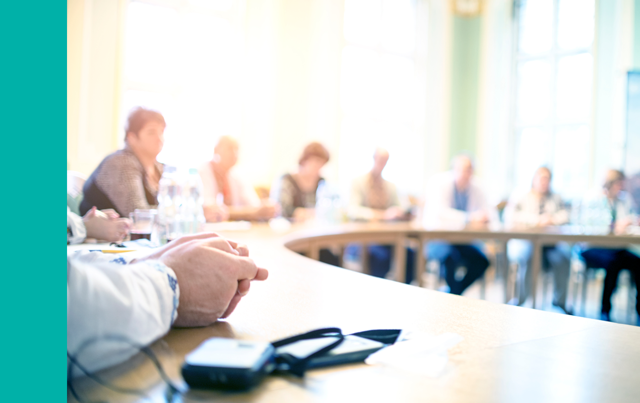 A group of people sits around a large, curved conference table in a bright meeting room with papers and a phone in the foreground.