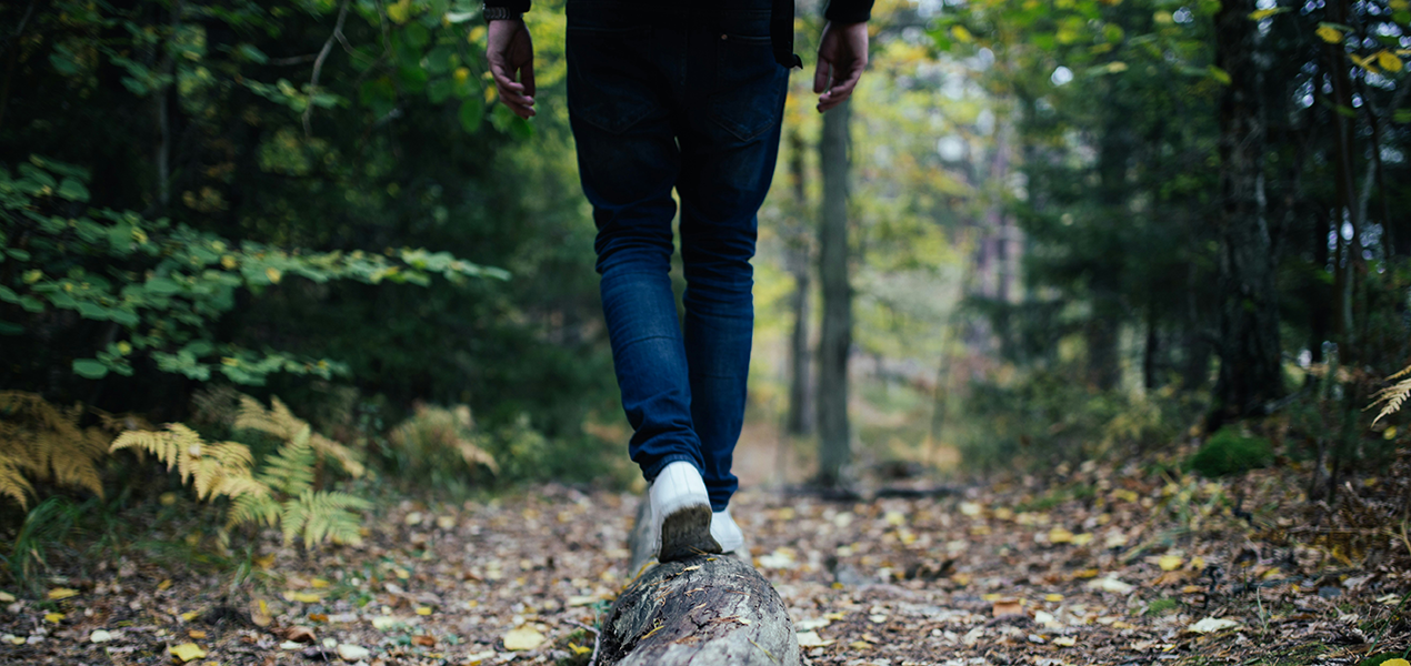 A person wearing blue jeans and white sneakers walks along a fallen log in a forest.