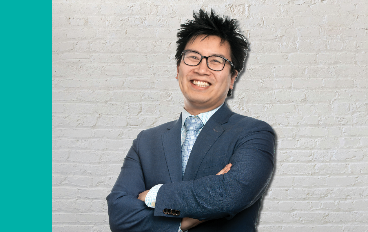 Chris Samuel, an Asian Man with black spiky hair, wearing glasses and a blue suit, poses in front of a light brick wall.