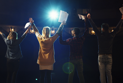 A group of people standing outdoors at night with their arms raised in celebration, illuminated by bright stage lights.