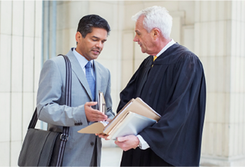 A male lawyer wearing a grey suit speaks to a male judge with grey hair wearing robes.