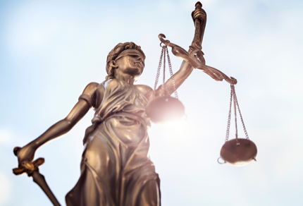 Bronze statue of Lady Justice holding a sword and balance scales, with sunlight shining in the background.