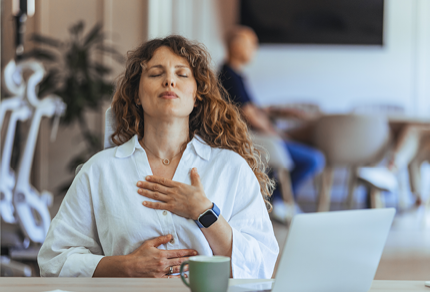 A woman sits at a desk with her eyes closed, one hand on her chest and the other on her abdomen, appearing to practice deep breathing. A laptop and mug are on the desk.