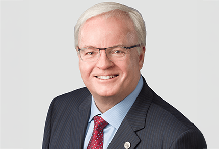 Tracey Stock, and man with short white hair and glasses wearing a dark pinstripe suit, light blue shirt, and red patterned tie, poses against a plain light background.