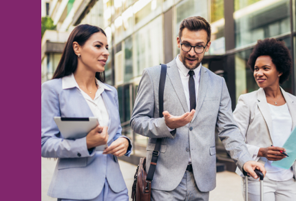 Three business professionals in suits walk and talk outside an office building; one holds a tablet, another gestures while speaking, and the third pulls a suitcase.