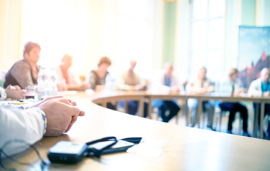 A group of people sit around a large conference table in a bright room, with documents and microphones on the table.