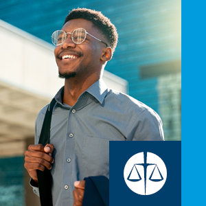 A professional Black man wearing glasses and a button-down shirt smiles while walking outdoors in an urban area on a sunny day.