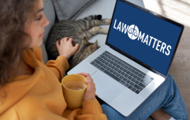 A person holding a mug sits on a couch next to a tabby cat, using a laptop with a blank white screen.