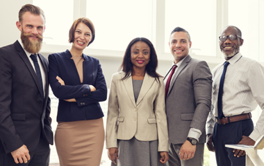 Five professionally dressed people stand and smile together in a modern, bright office space with large windows and a staircase.